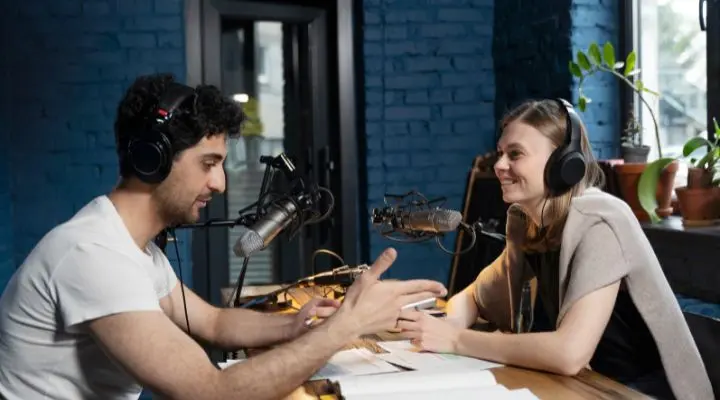 Dos personas con auriculares se sientan en una mesa con micrófonos, papeles y una computadora portátil, grabando un B2B podcast  en un estudio con paredes azules y plantas en el alféizar de la ventana.