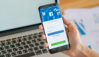 Una mano que sostiene un teléfono inteligente que muestra la pantalla de inicio de sesión de Facebook, con una computadora portátil abierta y documentos en el fondo.