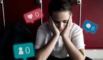 Un niño triste se sienta con la cabeza entre las manos, rodeado de íconos de redes sociales que muestran cero “me gusta”, comentarios y seguidores, frente a casilleros rojos.