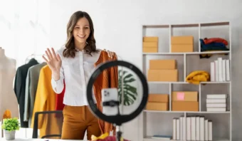 Una mujer, involucrada en el marketing de influencia, está parada frente a un teléfono inteligente y un anillo de luz, saludando y sosteniendo ropa, con estantes de cajas y artículos doblados en el fondo.