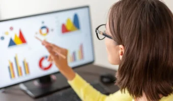Una mujer con gafas mira un monitor de computadora que muestra varios gráficos y cuadros de datos coloridos relacionados con la medición del marketing, mientras señala la pantalla con un bolígrafo.