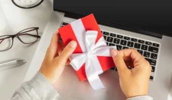 Manos sosteniendo un regalo (una pequeña caja de regalo roja con una cinta blanca) apoyado sobre el teclado de una computadora portátil, con gafas, un bolígrafo y un reloj cerca.