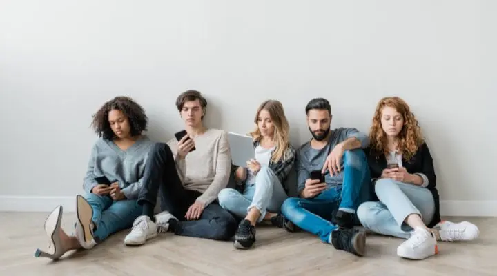 Cinco jóvenes adultos se sientan en el suelo contra una pared, cada uno absorto en su teléfono o tableta, navegando por las redes sociales y totalmente concentrados en sus dispositivos.