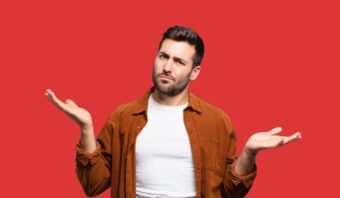 Un hombre con pelo corto y barba, de pie sobre un fondo rojo, levanta ambas manos con expresión desconcertada, como si se preguntara: ¿Assisted o Direct Commerce? Descubre cómo elegir el mejor modelo para conectar con tus clientes.