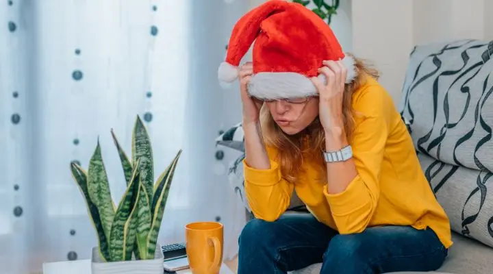 Una persona con gorro de Papá Noel y suéter amarillo está sentada en un sofá mirando hacia abajo con las manos en la cabeza, reflexionando sobre mi Navidad. Una planta, una taza y un control remoto están en la mesa cercana.