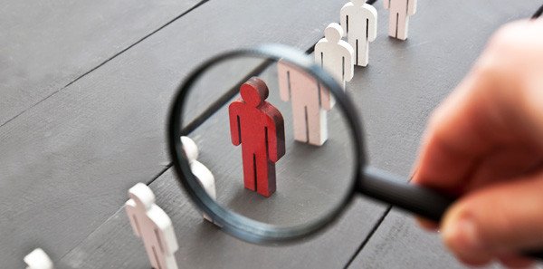 Una mano sostiene una lupa sobre una figura humana roja entre figuras blancas, lo que simboliza la selección o el reclutamiento, muy parecido a descubrir ¿qué es stalkear? donde se requiere un escrutinio minucioso para encontrar detalles específicos entre muchos.