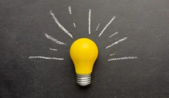 Una bombilla amarilla se encuentra sobre una superficie negra, con líneas de tiza blanca que irradian para simbolizar una idea brillante o inspiración: creatividad que no se puede robar.