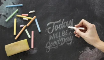Una mano escribe “Hoy será un buen día” con tiza en una pizarra, con tizas de colores y un borrador al lado de la escritura: inspiración perfecta para marketing en pizarrones.