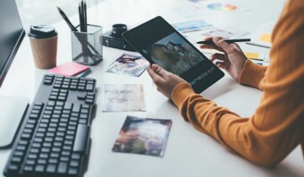 En un escritorio desordenado, adornado con un teclado, una taza de café e imágenes dispersas, alguien edita hábilmente fotos en una tableta, preparándolas para anuncios.