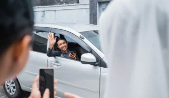 Un hombre sentado en un automóvil saluda y sonríe a dos personas afuera, una de las cuales sostiene un teléfono inteligente para tomar una foto o un video, capturando un momento amistoso de Uber México.