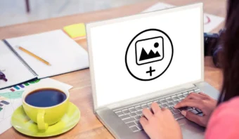 Persona escribiendo en una computadora portátil en un escritorio con una taza de café, papeles y una cámara; la pantalla de la computadora portátil muestra un ícono de carga de imágenes para GIF o Vídeo.