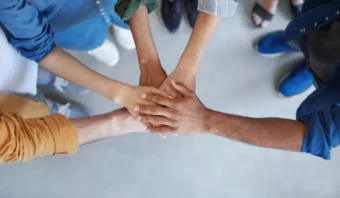 Un grupo de personas de pie en círculo con sus manos apiladas juntas en el centro, visto desde arriba, simboliza el poder del marketing colaborativo y el trabajo en equipo.
