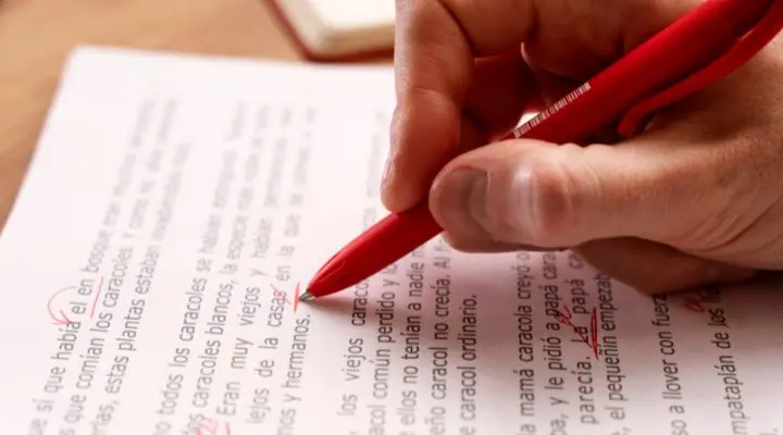 Una mano que sostiene un bolígrafo rojo realiza correcciones y marca ediciones en un documento impreso, centrándose en mejorar la ortografía.