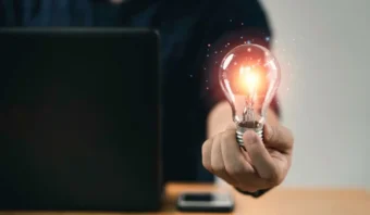 Una persona sostiene una bombilla encendida frente a una computadora portátil en un escritorio, con un teléfono inteligente al lado: listo para innovar y generar nuevas ideas.