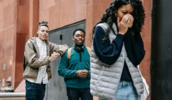 Una mujer se cubre el rostro y se aleja de dos hombres que están de pie contra una pared de ladrillos, aparentemente enfrascados en una conversación. La escena insinúa sutilmente la incomodidad del acoso laboral.