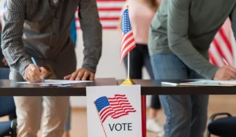 Dos personas llenan formularios en una mesa con una pequeña bandera estadounidense y un cartel que dice "VOTE", resaltando un evento de votación o registro a veces criticado por su poca efectividad.