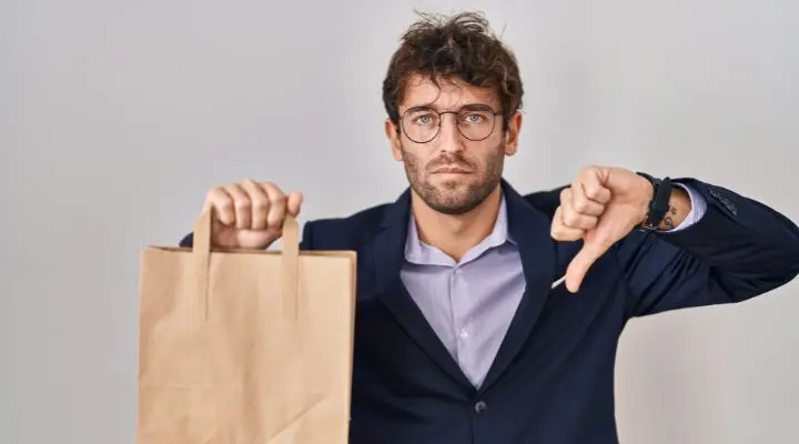 Un cliente insatisfecho, vestido de traje, sostiene en una mano una bolsa de papel marrón y con la otra hace un gesto de aprobación con el pulgar hacia abajo, de pie frente a un fondo liso.