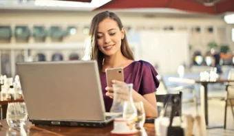 Una mujer se sienta en la mesa de un café al aire libre, sonriendo a su teléfono inteligente como una empresaria exitosa, con una computadora portátil abierta frente a ella y bebidas en la mesa.