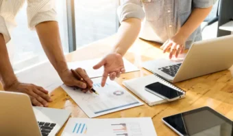 Dos personas trabajando en una mesa con computadoras portátiles, analizando gráficos y cuadros impresos mientras discuten la estrategia, con una libreta, un teléfono y una tableta también sobre la mesa.
