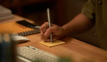Una persona escribe consejos prácticos para probar diseño gráfico en una nota adhesiva amarilla en un escritorio de madera cerca de un teclado de computadora y un cuaderno cerrado.