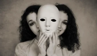 Dos mujeres están de pie juntas; una sostiene una máscara blanca sencilla con agujeros para los ojos frente a sus caras, ocultando parcialmente sus expresiones, un eco de la gente disfrazada de gente decente.