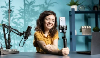 Una mujer se sienta en un escritorio con un micrófono, una computadora portátil y un teléfono sobre un trípode, aparentemente grabando o transmitiendo en vivo (quizás discutiendo el Síndrome de la Figura Pública) en una habitación con paredes azules y plantas.