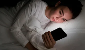 Persona acostada en la cama por la noche, iluminada por la luz de un teléfono inteligente, buscando la falsa felicidad mientras apoya su cabeza en una almohada.