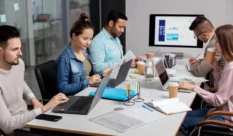 Cinco personas trabajan en una mesa con portátiles, papeles y tazas de café, manteniendo un ambiente de equipo concentrado. Un monitor al fondo muestra gráficos y tablas en esta moderna oficina o sala de reuniones, ideal para mantener a tu equipo concentrado.