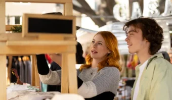 Dos personas se encuentran en un puesto de mercado al aire libre, una de ellas examina un artículo en un estante de madera que ofrece valor agregado, mientras la otra observa.