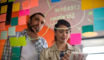 Dos personas están paradas frente a una pared de vidrio cubierta con notas adhesivas de colores y las palabras “Estrategia creativa”, mientras una mira una tableta, lista para probar sus ideas en una sesión de lluvia de ideas colaborativa.