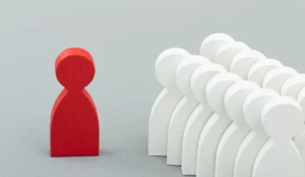 Una figura de madera roja se encuentra frente a un grupo de figuras de madera blanca dispuestas en filas sobre un fondo gris, resaltando el riesgo de lo exclusivo.