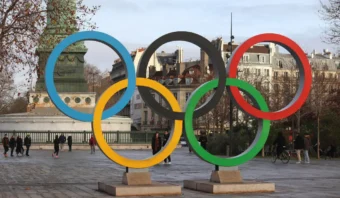 Una gran escultura de anillos olímpicos exhibida al aire libre en una plaza de la ciudad, con gente caminando y edificios históricos al fondo, mientras Internet rompe paradigmas en la transmisión de los Juegos Olímpicos alrededor del mundo.