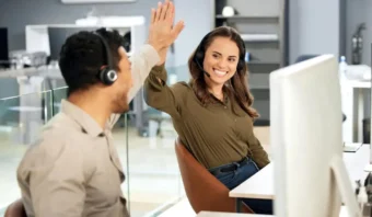 Dos trabajadores de oficina que usan auriculares sonríen y chocan los cinco mientras brindan un excelente servicio al cliente en un espacio de trabajo moderno.