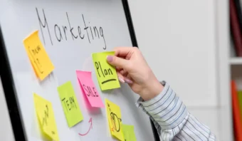 Una persona coloca una nota adhesiva rotulada "Plan" en una pizarra con "Marketing" en la parte superior, rodeada de notas coloridas, que ilustran los pasos de un plan de marketing en acción.