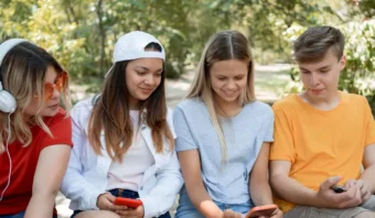Cuatro adolescentes están sentados juntos al aire libre; dos miran sus teléfonos para consultar las redes sociales mientras los otros dos observan, todos rodeados de árboles en un parque. "Estamos chupando tranquilos".