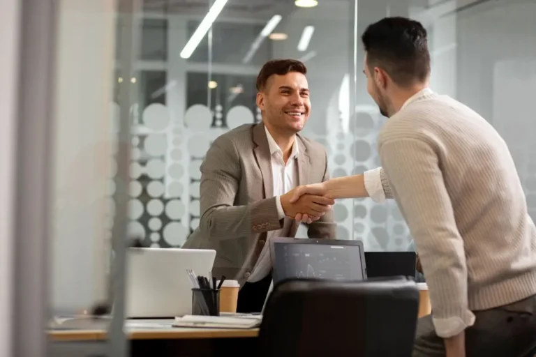 Dos hombres vestidos de negocios se dan la mano en un escritorio en una oficina moderna, ilustrando la dinámica de satisfacción vs. lealtad mientras discuten documentos y computadoras portátiles visibles en primer plano.