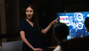 Una mujer se pone de pie y señala un monitor que muestra gráficos y cuadros coloridos, analizando estrategias fallidas en la política mientras presenta datos a colegas sentados en una oficina con poca luz.