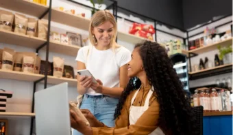 Dos mujeres en una tienda, una sosteniendo un teléfono inteligente y la otra usando una computadora portátil, sonriéndose cerca de estantes con productos empaquetados, mostrando cómo las pymes en redes sociales pueden conectarse y crecer en el mundo digital.