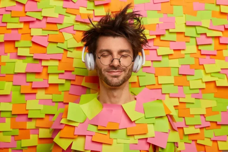Un hombre con el pelo revuelto y auriculares, con los ojos cerrados, está de pie frente a una pared llena de notas adhesivas de colores, con una camiseta hecha con notas adhesivas. ¿Te consideras creativo? Esta escena captura a la perfección la energía imaginativa.