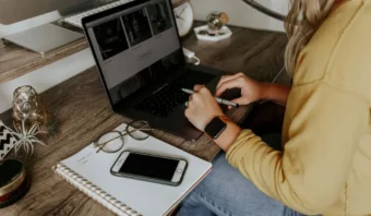 Una persona con un suéter amarillo usa una computadora portátil en un escritorio de madera, planificando campañas de mailing, con un cuaderno, un bolígrafo, un teléfono inteligente y gafas cerca.