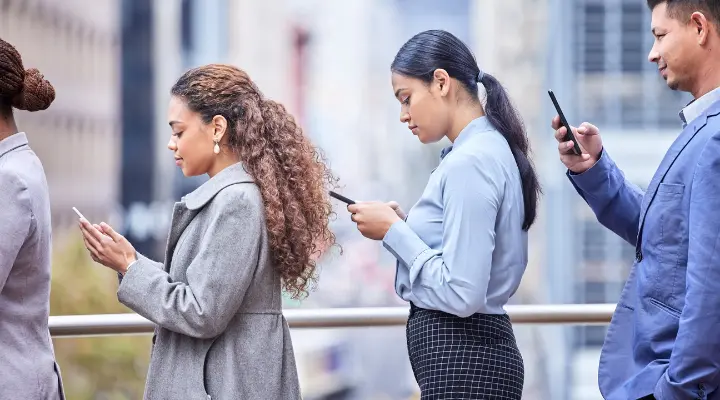 Cuatro personas forman fila al aire libre, cada una absorta en su teléfono inteligente frente a un paisaje urbano de fondo, una escena que recuerda a Black Mirror.
