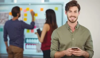 Un hombre con una camisa verde, posiblemente uno de los brand managers, sostiene un teléfono inteligente y sonríe a la cámara mientras dos personas trabajan con notas adhesivas en una pizarra en el fondo.