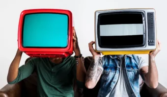 Dos personas se sientan en un sofá con televisores antiguos sobre sus cabezas —uno rojo, otro amarillo— frente a un fondo blanco liso, encarnando el encanto peculiar de las coincidencias.