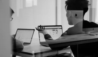 Dos personas trabajan en una mesa con portátiles, enfrascadas en una conversación sobre la entrevista que se muestra en sus pantallas. La imagen en blanco y negro se captura a través de una mampara de cristal.