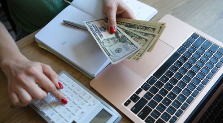 Una persona administra un presupuesto limitado, sosteniendo billetes de dólares estadounidenses mientras usa una calculadora, con un cuaderno y una computadora portátil abierta en el escritorio.