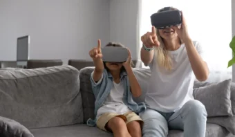 Un adulto y un niño se sientan en un sofá gris con cascos de realidad virtual, inmersos en una experiencia que da vida a lo virtual mientras ambos apuntan hacia adelante e interactúan con el mundo de realidad virtual.