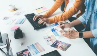 Dos personas en un escritorio trabajan con fotografías digitales e impresas. Una utiliza un teclado mientras la otra señala una copia impresa. Sobre el escritorio se ven una variedad de fotografías, una cámara y elementos para campañas impresas.