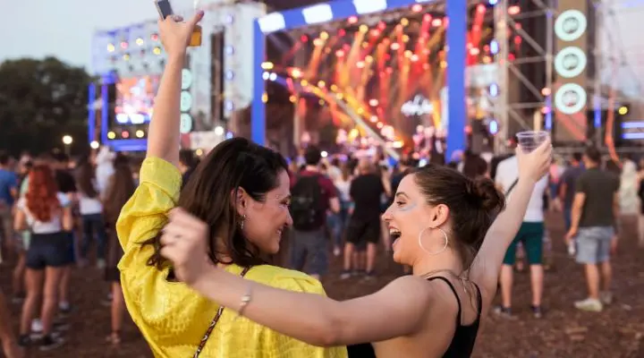 Dos personas en un concierto al aire libre animan con bebidas en la mano, rodeadas de una multitud. El escenario está iluminado con luces de colores. Festival AMAPRO 2019