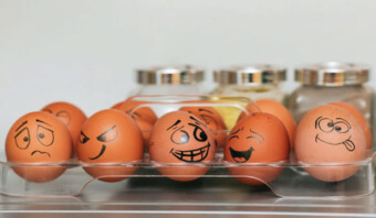 Huevos marrones en una bandeja transparente, cada uno con una cara de dibujos animados única dibujada en ellos, se colocan en un estante del refrigerador frente a frascos con tapas plateadas, creando una escena perfecta para ideas de publicidad emocional.