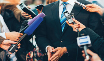 Una persona con traje está rodeada de periodistas que sostienen micrófonos y dispositivos de grabación durante una entrevista de prensa como parte de una campaña de Marketing Político.